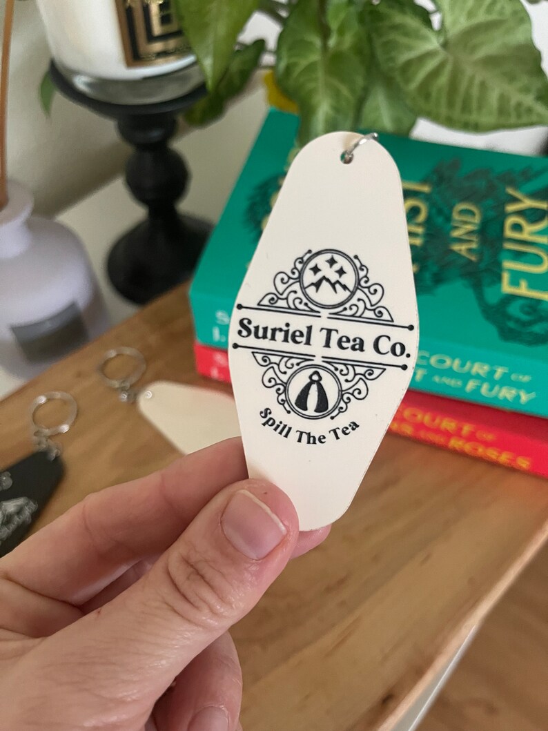 May include: White motel key tag with black text and a logo. The text reads "Suriel Tea Co. Spill The Tea".