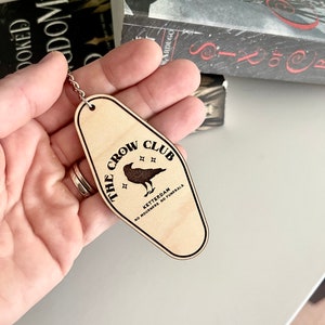 Peut inclure: Un porte-cl&eacute;s en bois en forme de cl&eacute; de motel avec le texte "The Crow Club" et "Ketterdam, No Mourners. No Funerals."  Le porte-cl&eacute;s pr&eacute;sente une illustration de corbeau noir.