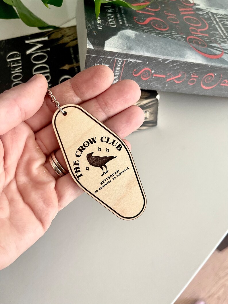 Peut inclure: Un porte-cl&eacute;s en bois en forme de cl&eacute; de motel avec le texte "The Crow Club" et "Ketterdam, No Mourners. No Funerals."  Le porte-cl&eacute;s pr&eacute;sente une illustration de corbeau noir.