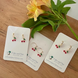 May include: Three pairs of cherry earrings on white cards with the text "Palm and Pine Design 321" and the website address "palmandpine32L.etsy.com". One pair is a hoop earring with a cherry dangling from it. The other two pairs are dangle earrings with a cherry charm.