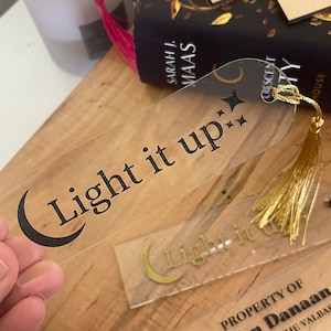 Op de afbeelding: Een transparante acryl bladwijzer met een zwarte halve maan en de tekst "Light it up" in zwarte letters. De bladwijzer heeft een gouden kwastje.