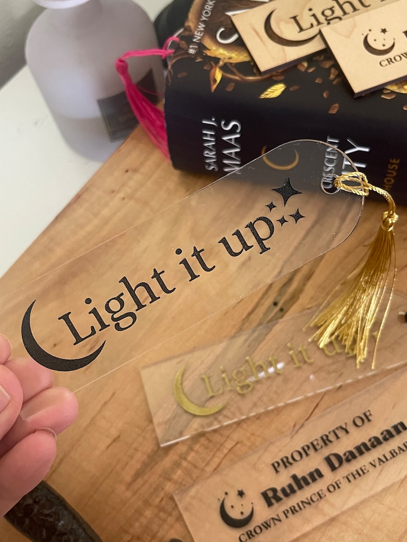 Op de afbeelding: Een transparante acryl bladwijzer met een zwarte halve maan en de tekst "Light it up" in zwarte letters. De bladwijzer heeft een gouden kwastje.