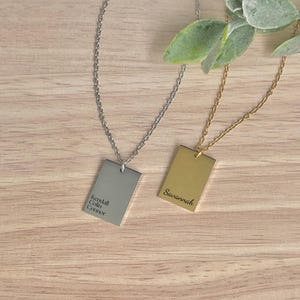 May include: Two rectangular pendant necklaces. One is silver with the names "Kendall, Colin, Connor" engraved. The other is gold with the name "Savannah" engraved. Both necklaces hang on delicate chains, set against a wooden background.
