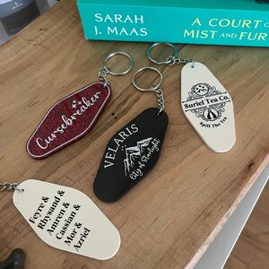 May include: Four keychains with different designs. One is red with the text "Cursebreaker" in white. Another is black with the text "VELARIS City of Starlight" in white. A third is white with the text "Feyre & Rhysand & Amren & Cassian & Mor & Azriel" in black. The last is white with the text "Suriel Tea Co. Spill The Tea" in black.