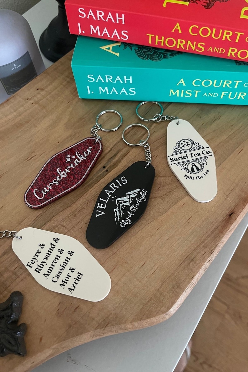 May include: Four keychains with different designs. One is red with the text "Cursebreaker" in white. Another is black with the text "VELARIS City of Starlight" in white. A third is white with the text "Feyre & Rhysand & Amren & Cassian & Mor & Azriel" in black. The last is white with the text "Suriel Tea Co. Spill The Tea" in black.