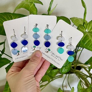 May include: Three pairs of dangle earrings in shades of blue, teal, and white are displayed. Each earring features a series of geometric shapes, including circles and ovals, suspended from a silver hook. The earrings are presented on white cards with the text "Palm and Pine Design 321".