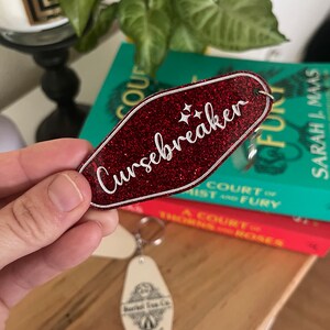 May include: Red glitter keychain with a white outline and the text "Cursebreaker" in white lettering. The keychain is shaped like a motel key.