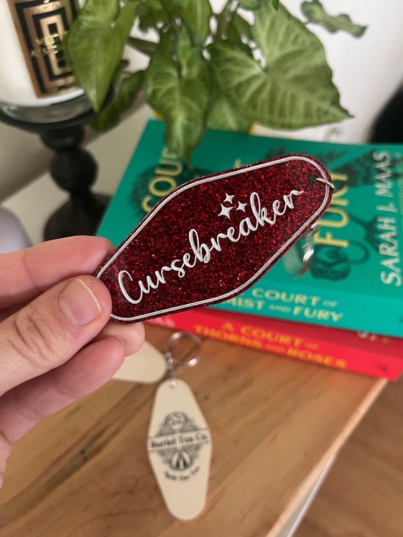 May include: Red glitter keychain with a white outline and the text "Cursebreaker" in white lettering. The keychain is shaped like a motel key.
