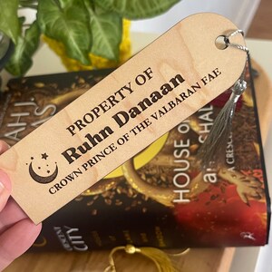 Op de afbeelding: Een houten bladwijzer met een halve maan en sterren ontwerp. De bladwijzer is gegraveerd met de tekst "PROPERTY OF Ruhn Danaan CROWN PRINCE OF THE VALBARAN FAE".