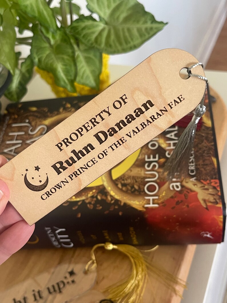 Op de afbeelding: Een houten bladwijzer met een halve maan en sterren ontwerp. De bladwijzer is gegraveerd met de tekst "PROPERTY OF Ruhn Danaan CROWN PRINCE OF THE VALBARAN FAE".