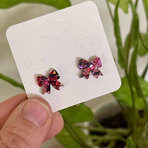 May include: A pair of red and pink glitter bow earrings on a white card.