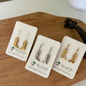 May include: Three pairs of cowboy boot earrings in gold and silver tones, displayed on white cards. The earrings are detailed with classic western boot designs. The cards have the text "Palm and Pine DESIGN 321 palmandpine32letsty.com".