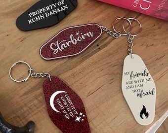 May include: A collection of keychains with various designs and text. One black keychain reads "PROPERTY OF RUHN DANAAN." A red glitter keychain says "Starborn." Another red keychain has a moon and stars with "LIGHT IT UP." A cream keychain says "My friends are with me and I am not afraid."