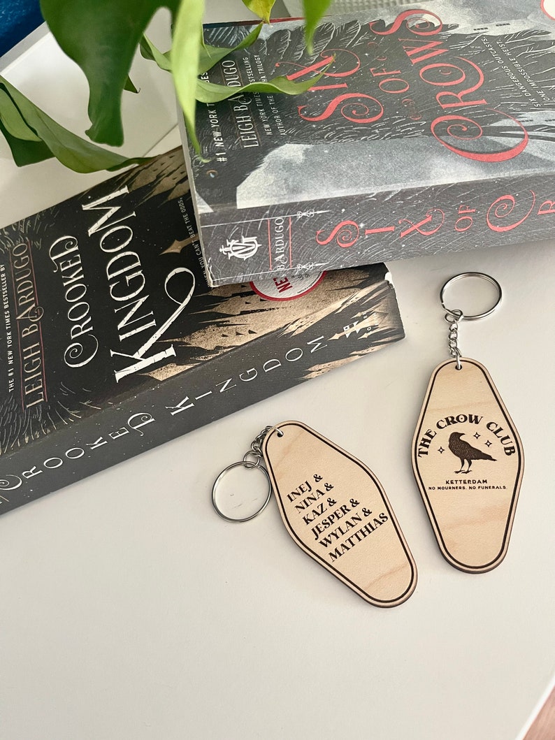Peut inclure: Deux porte-cl&eacute;s en bois avec un motif de corbeau et le texte "The Crow Club" et "Ketterdam No Mourners No Funerals". L'autre porte-cl&eacute;s porte le texte "Kaz & Nina & Inej & Matthias & Jesper & Wylan".