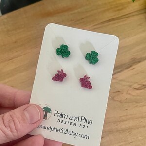 May include: A pair of earrings on a white card. The earrings are shaped like green glitter shamrocks and pink glitter bunnies. The card has the text "Palm and Pine DESIGN 321" and the website address "palmandpine321.etsy.com".