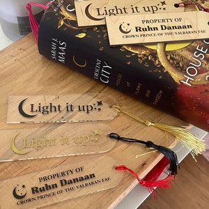 Op de afbeelding: Twee transparante acryl-bladwijzers met gouden letters die "Light it up" en "Property of Ruhn Danaan Crown Prince of the Valbaran Fae" zeggen. De bladwijzers hebben een halve maan en ster ontwerp.