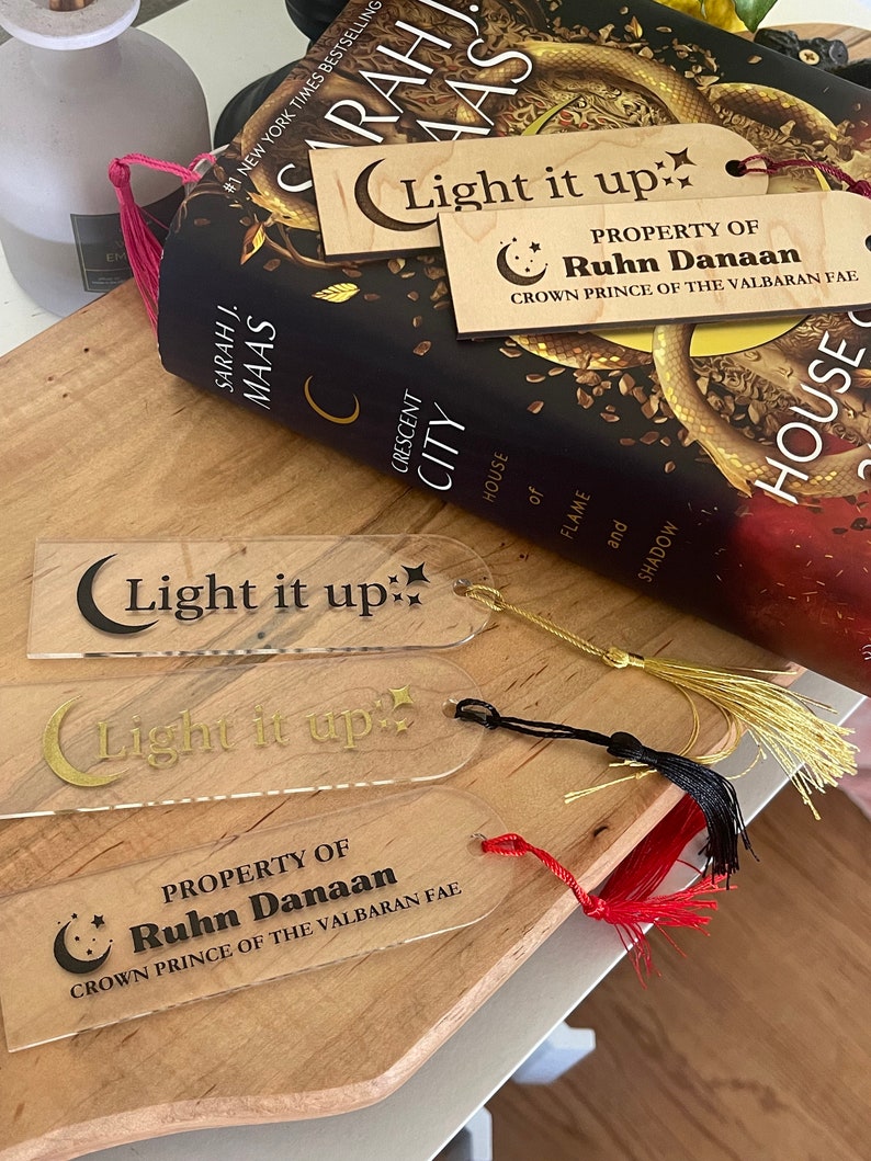 Op de afbeelding: Twee transparante acryl-bladwijzers met gouden letters die "Light it up" en "Property of Ruhn Danaan Crown Prince of the Valbaran Fae" zeggen. De bladwijzers hebben een halve maan en ster ontwerp.