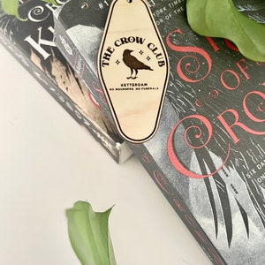 Peut inclure: Un porte-cl&eacute;s en bois en forme de cl&eacute; de motel avec le texte "The Crow Club" et une illustration de corbeau. Le texte "Ketterdam" et "No Mourners. No Funerals." sont &eacute;galement inclus sur le porte-cl&eacute;s.