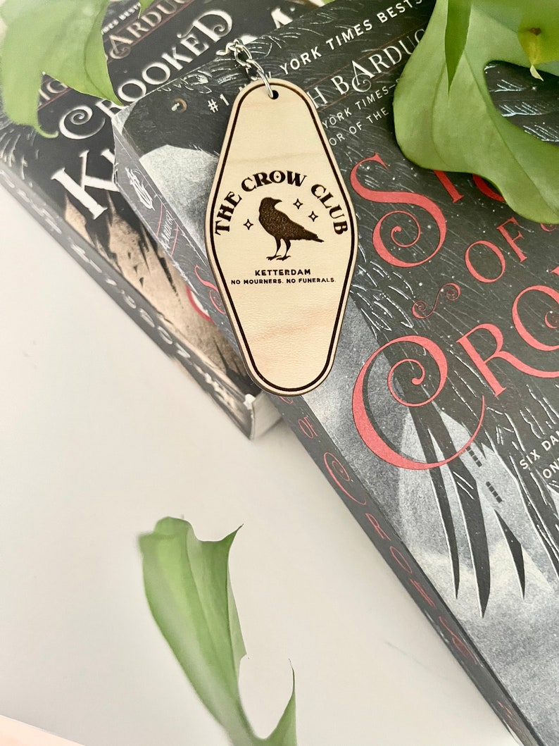 Peut inclure: Un porte-cl&eacute;s en bois en forme de cl&eacute; de motel avec le texte "The Crow Club" et une illustration de corbeau. Le texte "Ketterdam" et "No Mourners. No Funerals." sont &eacute;galement inclus sur le porte-cl&eacute;s.