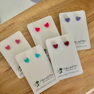 May include: Five pairs of heart-shaped earrings on white cards. The earrings are in various colors, including pink, red, purple, turquoise, and glitter. The cards have the text "Palm and Pine Design 321" and a website address.