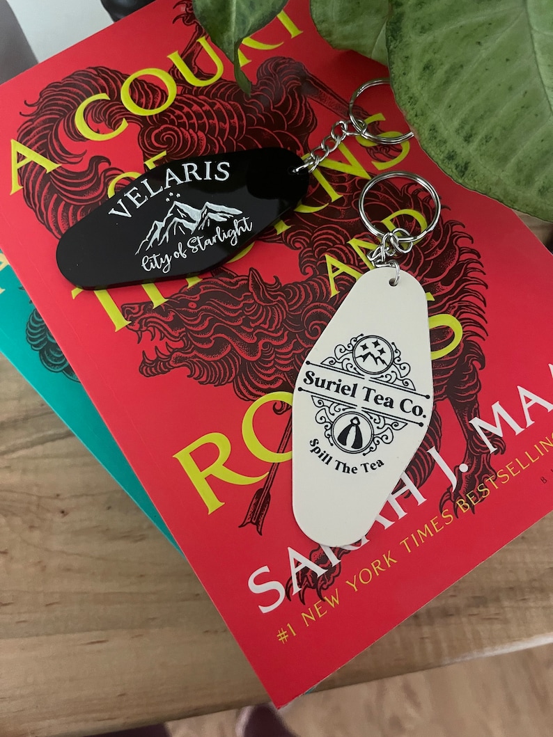 May include: Two keychains with black and white enamel designs. The black keychain features the text "Velaris City of Starlight" and a mountain range. The white keychain features the text "Suriel Tea Co. Spill The Tea" and a stylized teacup.
