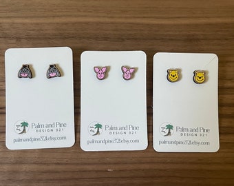 May include: Three pairs of wooden earrings featuring cartoon characters from Winnie the Pooh. The first pair features Eeyore, the second pair features Piglet, and the third pair features Winnie the Pooh. The earrings are on white cards with the text "Palm and Pine Design 321" and a website address.