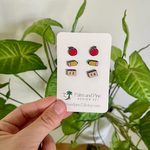 May include: A set of wooden stud earrings featuring school-themed designs: red apples, yellow pencils, and rulers. The earrings are displayed on a white card with the text "Palm and Pine Design 321" and a website address.