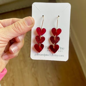 May include: Red glitter heart dangle earrings with gold hooks. The earrings are layered with three hearts, each with a different shade of red. The hearts are made of acrylic and have a shiny finish. The earrings are displayed on a white card with the text "Palmandpine321.etsy.com".