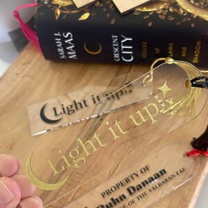Op de afbeelding: Een transparante acryl boekenlegger met gouden letters die "Light it up" lezen en een halve maan ontwerp. De boekenlegger heeft ook een zwart en rood kwastje. De boekenlegger is eigendom van Ruhn Danaan, Kroonprins van de Valbaran Fae.