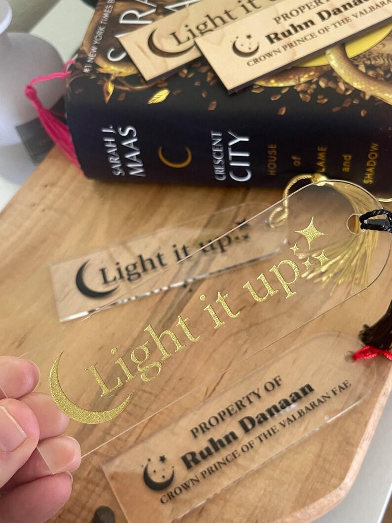 Op de afbeelding: Een transparante acryl boekenlegger met gouden letters die "Light it up" lezen en een halve maan ontwerp. De boekenlegger heeft ook een zwart en rood kwastje. De boekenlegger is eigendom van Ruhn Danaan, Kroonprins van de Valbaran Fae.