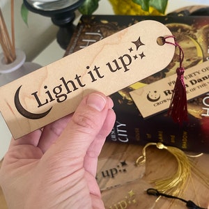 Op de afbeelding: Een houten bladwijzer met een halve maan en sterren ontwerp. De bladwijzer is gegraveerd met de tekst "Light it up:". De bladwijzer heeft een rode kwast.