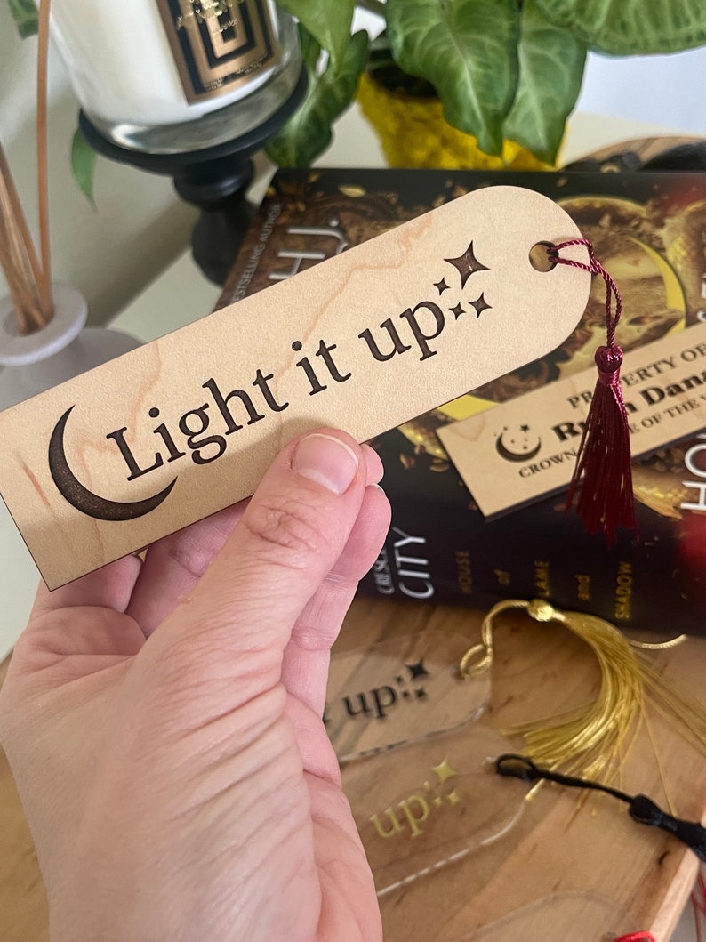 Op de afbeelding: Een houten bladwijzer met een halve maan en sterren ontwerp. De bladwijzer is gegraveerd met de tekst "Light it up:". De bladwijzer heeft een rode kwast.