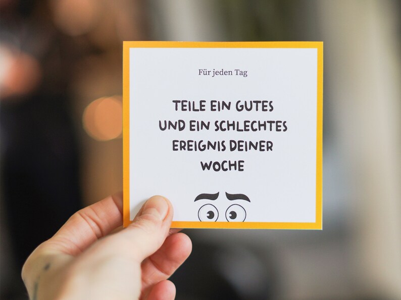 May include: A yellow and white card with the text "F&uuml;r jeden Tag" and "TEILE EIN GUTES UND EIN SCHLECHTES EREIGNIS DEINER WOCHE". The card has a cartoon face with eyebrows and eyes.