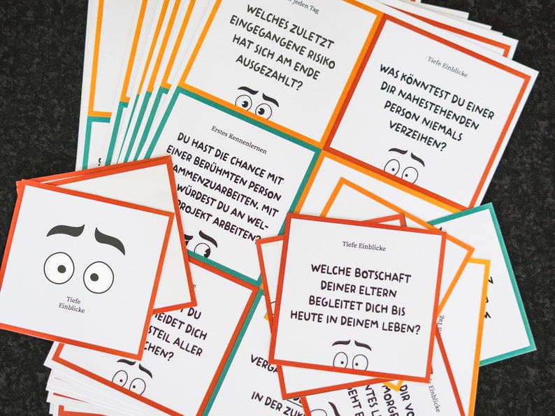 May include: A stack of white cards with orange, green, and red borders. Each card has a question printed on it in black text. The questions are about personal experiences and deep insights. The cards are designed for a game or activity.