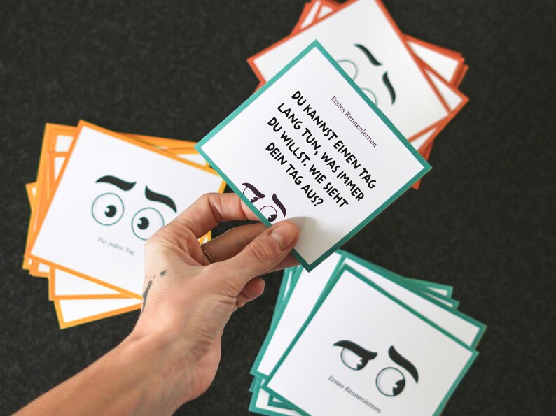 May include: A stack of cards with a green and white card on top. The card has the text "DU KANNST EINEN TAG LANG TUN, WAS IMMER DU WILLST. WIE SIEHT DEIN TAG AUS?" and the text "Erstes Kennenlernen".