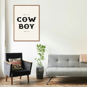 May include: A framed print with a white background and black text that reads "you're a COWBOY like me". The print is hanging on a white wall above a gray sofa and a black armchair.