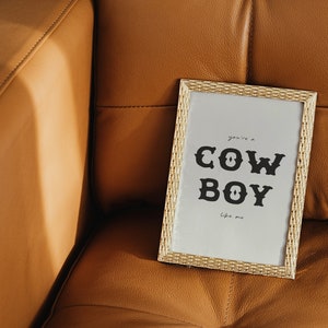 May include: A gold framed print with the text "you're a COWBOY like me" on a light brown leather couch.