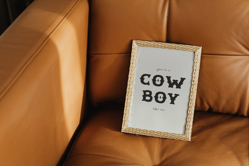 May include: A gold framed print with the text "you're a COWBOY like me" on a light brown leather couch.