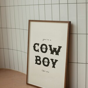 May include: A framed print with a white background and black text that reads "you're a COWBOY like me". The print is in a brown wooden frame.