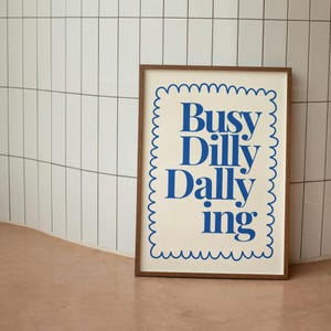 May include: A framed print with a white background and blue text that reads "Busy Dilly Dally ing" in a decorative font. The frame is brown wood with a scalloped border.