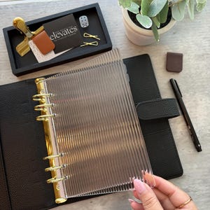 May include: A clear, ribbed planner divider is held over a black leather-bound planner with gold-colored rings. A black tray holds a gold clip, business cards with the word "elevates", a small brown leather card holder, and gold paper clips. A pen and a small brown leather bookmark are also visible.