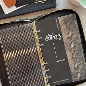 May include: Black zippered planner with gold accents, open to reveal a clear divider and pages with text that reads "my energy is expensive." A black tray holds business cards, a dice, and gold paper clips.