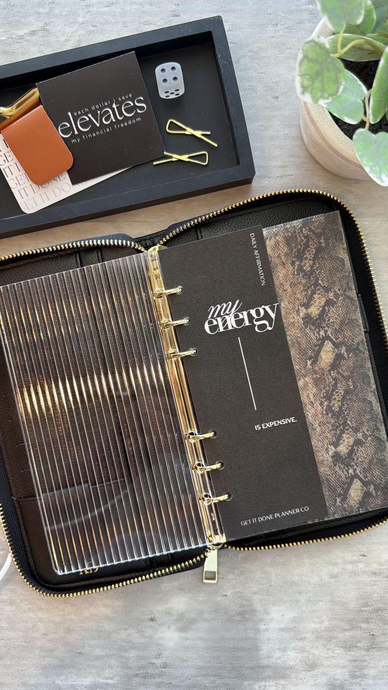 May include: Black zippered planner with gold accents, open to reveal a clear divider and pages with text that reads "my energy is expensive." A black tray holds business cards, a dice, and gold paper clips.