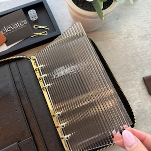 以下が含まれることがあります： ゴールドのジッパーと透明なリブ付きの仕切りが付いた黒革のバインダー。 バインダーは開いており、内部が明らかになっています。 アクセサリーが入った小さなトレイと鉢植えの植物が背景にあります。 「elevates」というテキストがカードに表示されています。