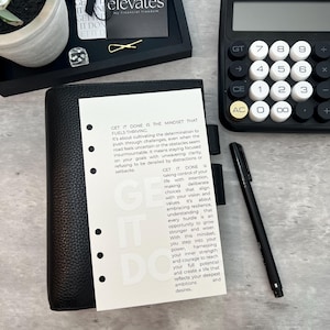 May include: A black leather planner with a white insert featuring the text "GET IT DONE." A black calculator, pen, and decorative tray with a gold paperclip are also visible. A small succulent plant in a white pot is in the background.