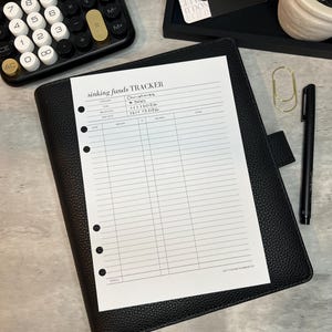 May include: A black leather planner with a "sinking funds tracker" sheet, a calculator, a pen, and a gold paper clip. The tracker sheet has the words "Christmas", "GOAL", "BEGIN DATE", and "END DATE" printed on it. A small black tray with a tag that says "elevates my financial freedom" is also visible.