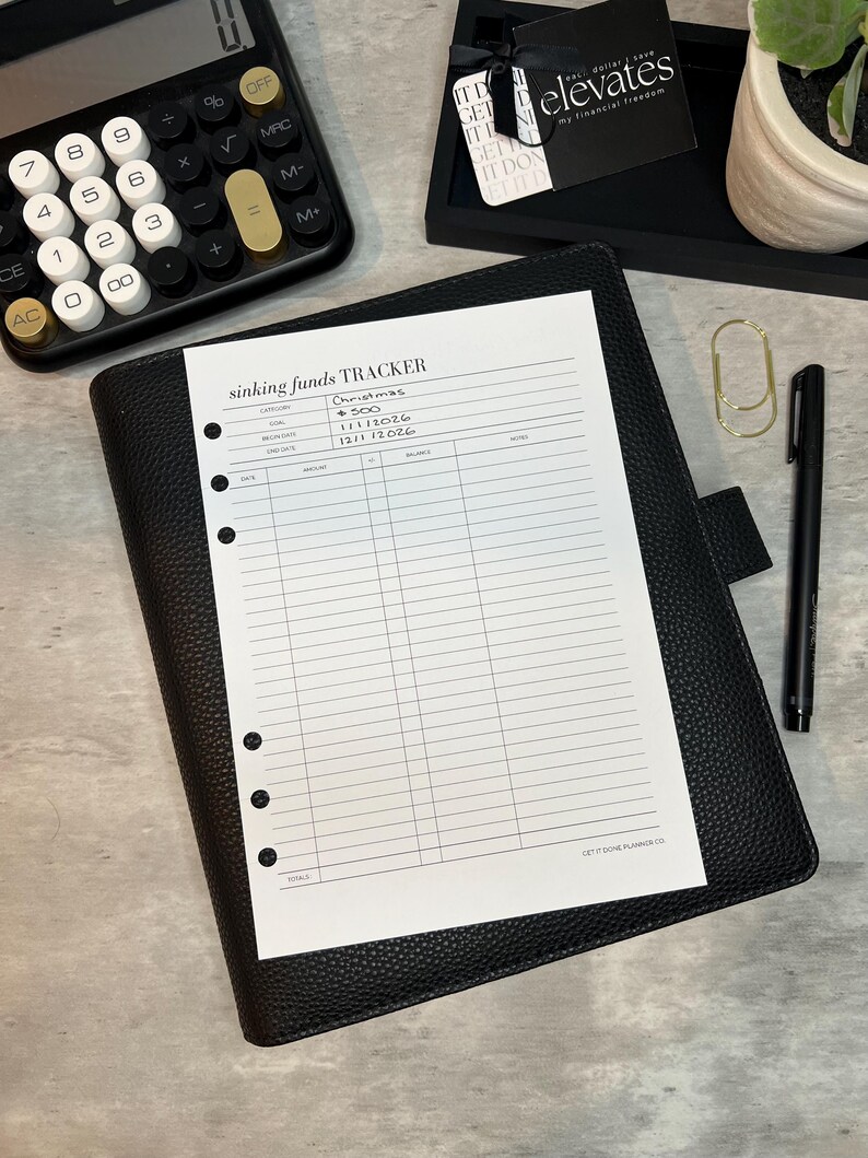 May include: A black leather planner with a "sinking funds tracker" sheet, a calculator, a pen, and a gold paper clip. The tracker sheet has the words "Christmas", "GOAL", "BEGIN DATE", and "END DATE" printed on it. A small black tray with a tag that says "elevates my financial freedom" is also visible.