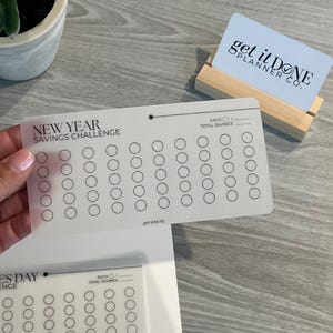 May include: A clear plastic sheet with a New Year Savings Challenge tracker. The sheet has 6 rows of 10 circles each. The text "EACH" and "TOTAL SAVINGS" are written at the top of the sheet. The text "get it DONE" is written at the bottom of the sheet.
