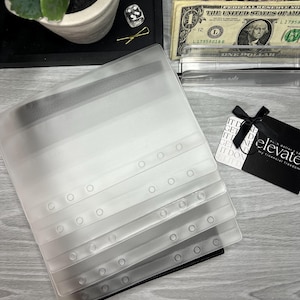 May include: A stack of clear, plastic binder sleeves with reinforced edges. The sleeves are partially transparent, with a gradient from white to black. A black tray holds a small plant, dice, and a gold paperclip. US currency and a gift card are also visible.