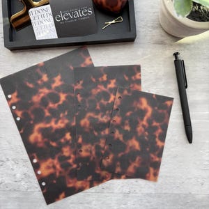 May include: A collection of stationery items, including tortoise shell patterned planner inserts, a black pen, and a black tray with a gold clip and business cards. The business cards read "elevates my financial freedom". An AirPod case with a tortoise shell pattern is also present.